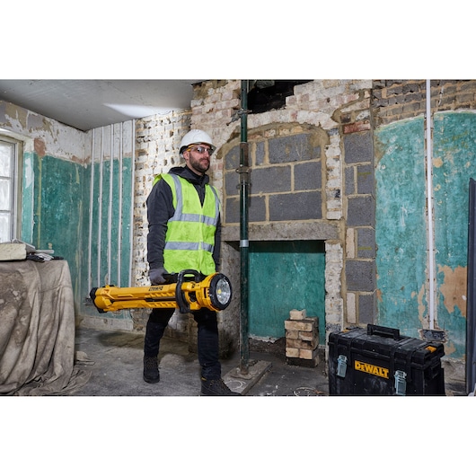 Worker in safety gear carries a DEWALT DCL079 light in a construction site, next to a DEWALT tool case.