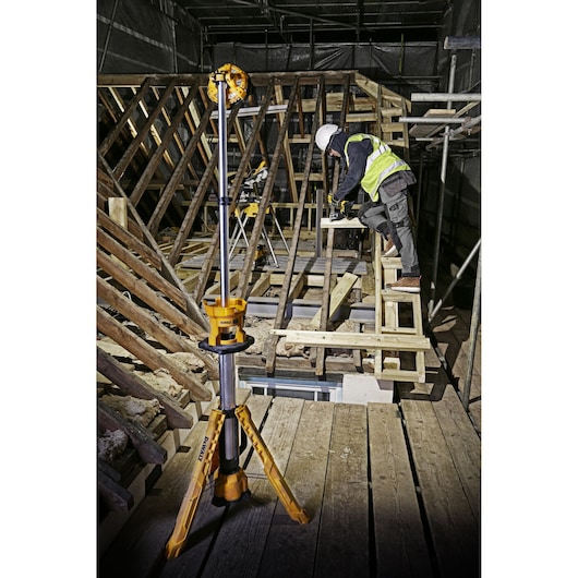 DEWALT DCL079 tripod work light is placed in a construction area where a worker is building wooden framework.