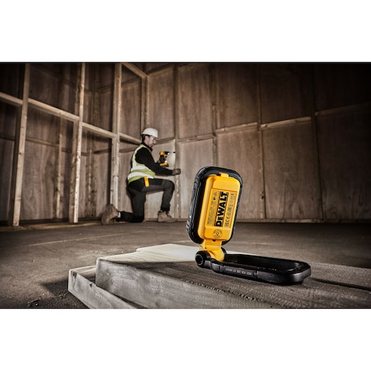 DEWALT DCL182 portable work light placed on a construction site floor, with a person in safety gear kneeling in the