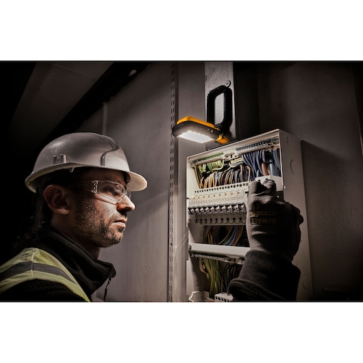 A worker wearing a hard hat and gloves is inspecting or working on an electrical panel, which is illuminated by a DEWALT