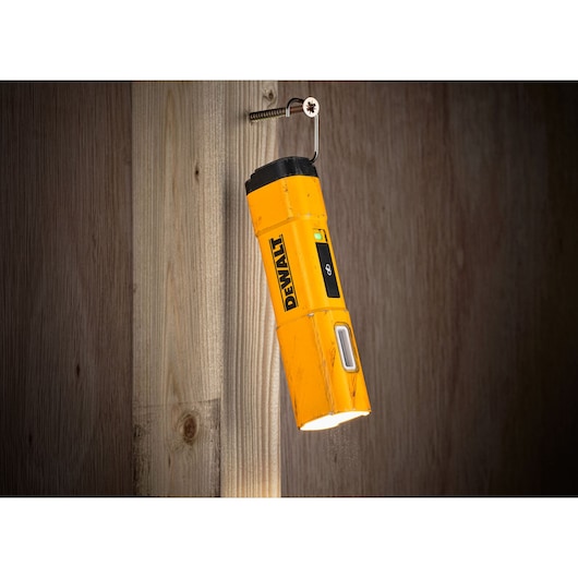 A yellow DEWALT DCL183 portable LED flashlight is hanging from a hook screwed into a wooden wall, illuminating downward.