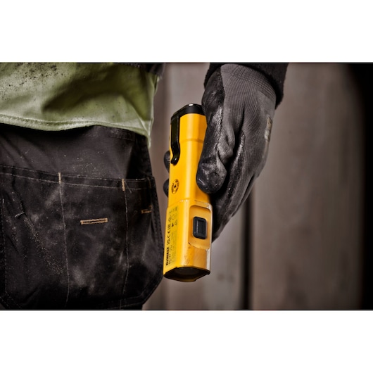 A person wearing black work gloves and work pants holds a yellow DEWALT DCL183 flashlight at their side.
