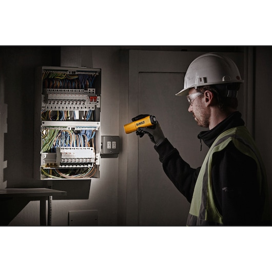 A person wearing a safety helmet and vest uses a DEWALT handheld flashlight to inspect an electrical panel with multiple