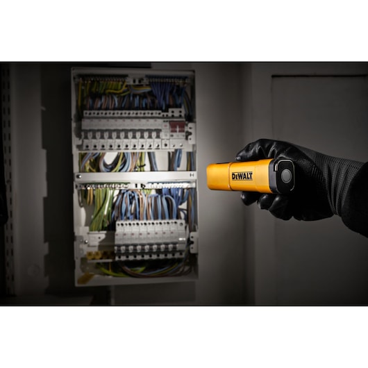 A person wearing a black glove is holding a yellow DEWALT flashlight, shining it towards an electrical panel with multiple