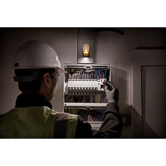 A worker wearing a safety helmet, reflective vest, and gloves is inspecting an electrical control panel with multiple wires