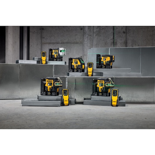 A group of DEWALT laser level and detector tools, arranged in a staged industrial setting with concrete walls and metallic platforms. The devices feature yellow and black designs with visible green laser lines.
