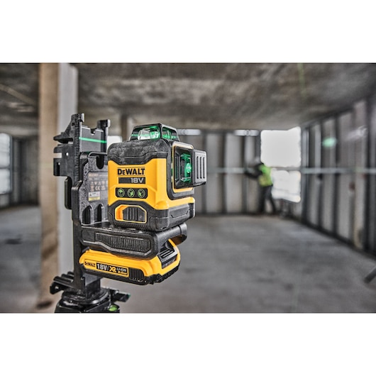 A DEWALT 18V laser level device, model DCLE34031D1, is mounted on a tripod in an unfinished building interior. The green laser lines are visible, and a worker in safety clothing is in the background aligning materials for construction.