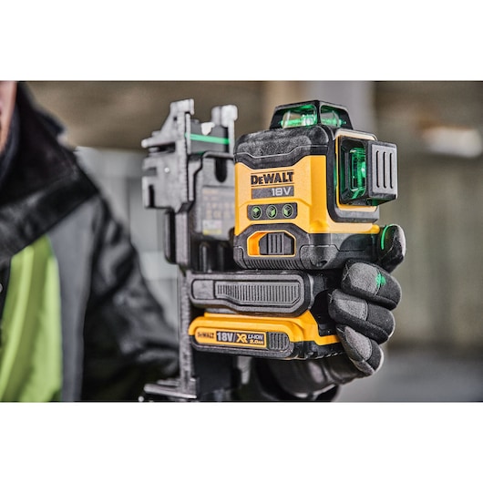 A close-up view of a person holding a DEWALT 18V laser level, used for precise measurements in construction and photography. The device features green laser lights and an XR lithium-ion battery.