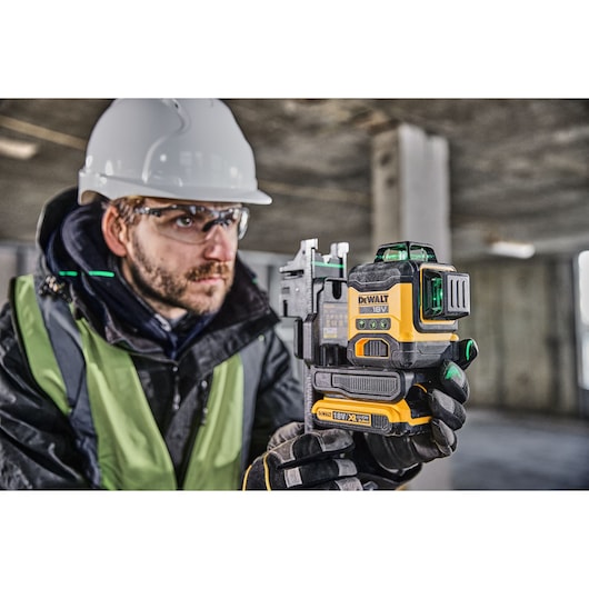 A person wearing a safety helmet and reflective vest is holding a DEWALT 18V green laser level tool (model DCLE34031N) at a construction site.