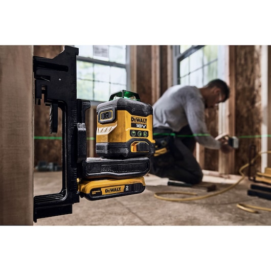 DEWALT 18V XR green line laser level mounted on a bracket with battery attached, being used on a construction site. A person is working in the background with blurred face, and the green laser line is visible across the workspace.