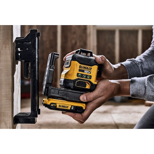 A close-up of hands positioning a DEWALT 18V laser level device, model DCLE34031N, against a vertical surface inside a construction site. The device is yellow and black with clear labeling and battery compartment visible.