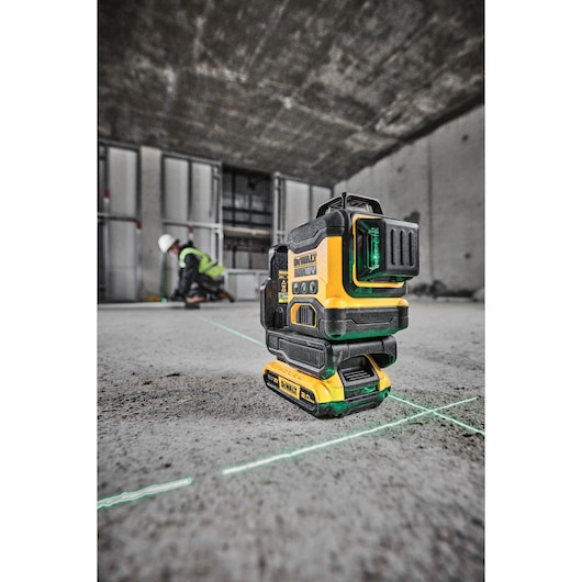 A DEWALT 18V green line laser level placed on the floor of a construction site, projecting green laser lines. In the background, a worker wearing a safety helmet and vest is working near a wall.