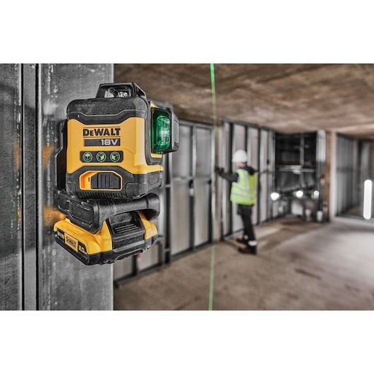 A DEWALT 18V green laser level is attached to a metal frame in a construction site. In the background, a person wearing a high-visibility vest and hard hat is working on the wall structure, with the green laser line used for alignment.