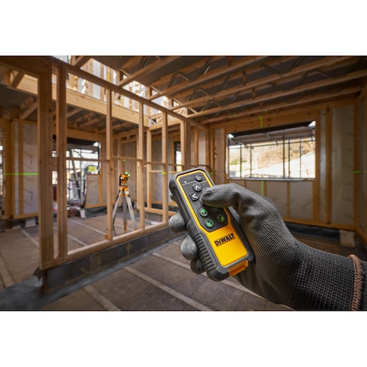 Gloved hand holds a DEWALT remote in a wooden-framed construction site. Laser level device visible in the background.