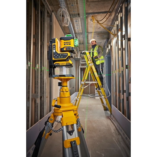 DEWALT laser level on tripod in a construction site, worker on ladder using green laser lines for alignment.