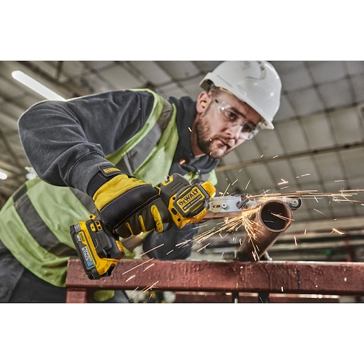 Worker uses DEWALT cordless tool to cut metal pipe, producing sparks. Indoor industrial setting, safety gear visible.