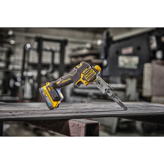 A DEWALT cordless electric file tool with a yellow and black design is placed on a metal surface in an industrial workshop setting.