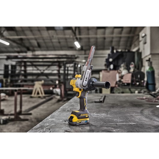 DEWALT DCM200E2T cordless power tool with a sanding belt attachment placed on a worktable in an industrial workshop setting.