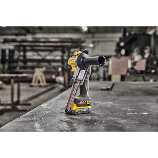 A DEWALT cordless handheld power tool, specifically a belt sander, is standing upright on a workshop table. The tool features a yellow and black design, a side handle, and a belt sanding mechanism in the front. The background shows a blurred industrial workspace with equipment and structures.
