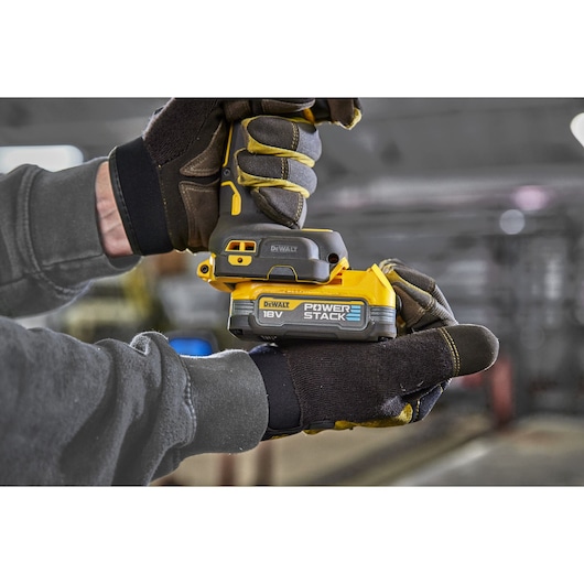 Gloved hands hold a DEWALT 18V Powerstack battery and tool in an industrial setting.