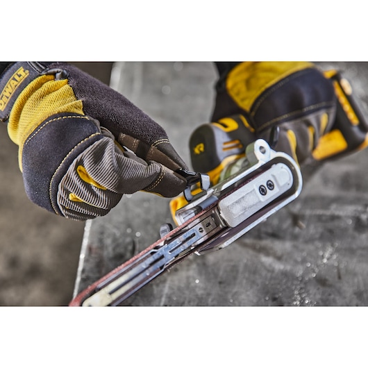 Gloved hand adjusts a DEWALT belt sander tool on a work surface. Tool has yellow and black accents.