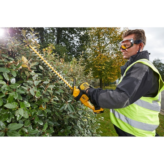 Person trimming hedge with yellow DEWALT cordless hedge trimmer outdoors, wearing gloves and safety vest.
