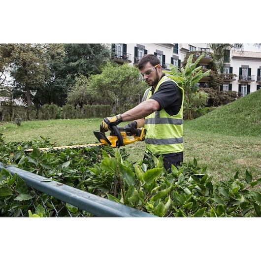 Man trims hedge with DEWALT DCM563PB cordless hedge trimmer in a garden, wearing gloves and safety vest.