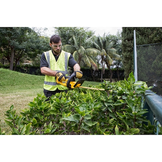 Worker trims hedge with yellow DEWALT DCM563PB tool outdoors, wearing gloves and safety vest.