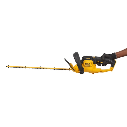 A DEWALT DCM563PB cordless hedge trimmer being held in a gloved hand, featuring a yellow and black design with a long blade.