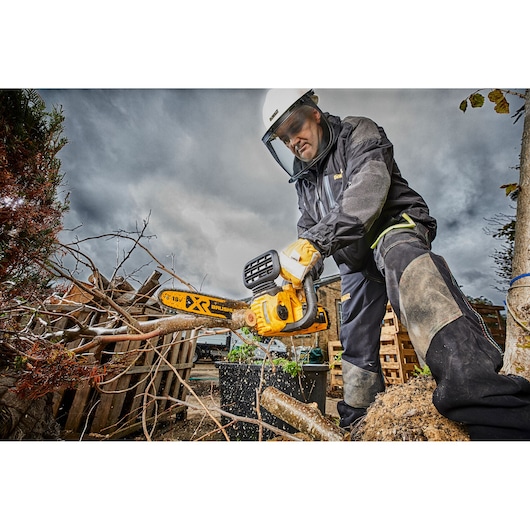 Person using a DEWALT cordless chainsaw to cut tree branches outdoors, wearing protective gear.