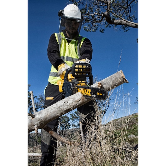 DEWALT DCM565P1 chainsaw cutting a branch outdoors. Person wearing safety gear and hi-vis vest.