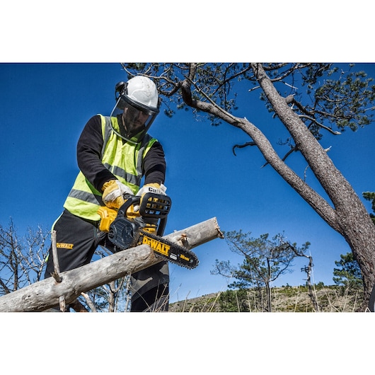 Person using a DEWALT chainsaw to cut a tree branch outdoors, wearing safety gear and a reflective vest.
