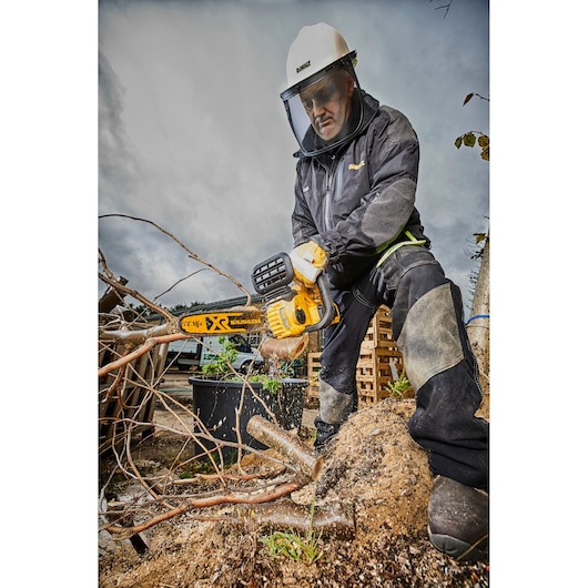 Person in safety gear uses a DEWALT chainsaw outdoors, cutting tree branches. Sawdust is visible.