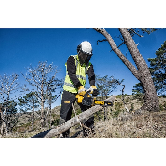 Person using a DEWALT DCM565P1 chainsaw outdoors, wearing safety gear and cutting a tree branch.