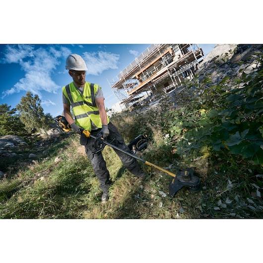 Worker uses DEWALT brush cutter outdoors, wearing safety gear near a construction site with scaffolding.