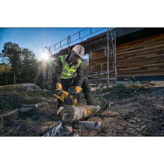 Worker uses a DEWALT chainsaw to cut logs outdoors, wearing safety gear near a wooden building and scaffold.