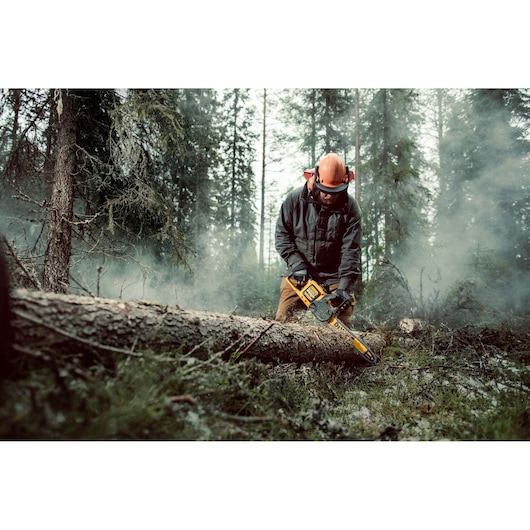 Person using a DEWALT chainsaw to cut a fallen tree in a misty forest. Worker wears protective gear.