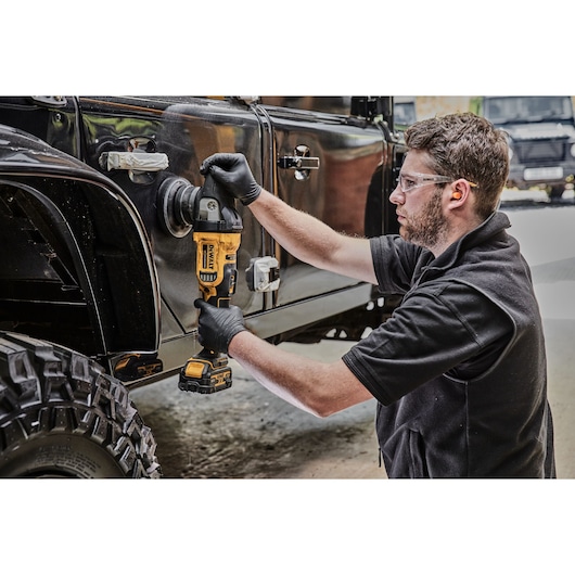 A person wearing black gloves uses a DEWALT cordless polisher (model DCM848N) to polish the side of a black off-road vehicle. The scene takes place in a workshop or garage.