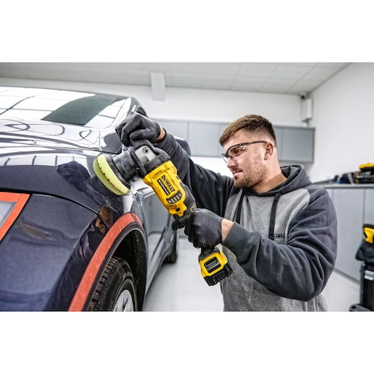 Person uses DEWALT cordless polisher DCM848P2 to polish a car in a workshop. Face is blurred for privacy.