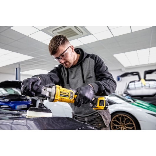 Person using DEWALT DCM849P2 polisher on car surface in a modern garage with luxury vehicles in the background.