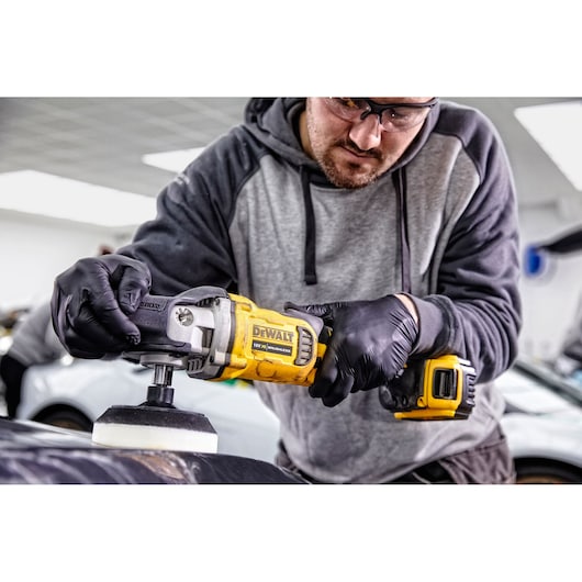 Person using a DEWALT DCM849P2 cordless polisher to work on a car surface in a garage, wearing gloves.
