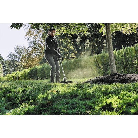 DEWALT DCZ65713X1-XE, DCMAS5713N in use outdoors trimming grass near a tree.