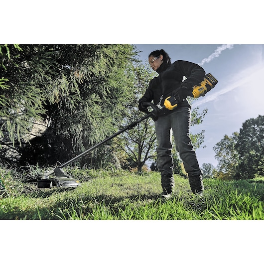 Person using a DEWALT cordless grass trimmer outdoors on a lawn with trees in the background.