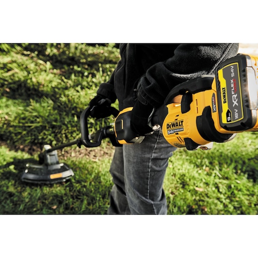 Person uses a DEWALT 54V cordless brushless grass trimmer outdoors in a garden.