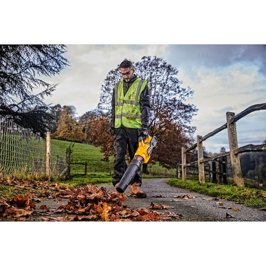 Person uses a DEWALT leaf blower on a rural path, wearing a safety vest, with autumn leaves on the ground.