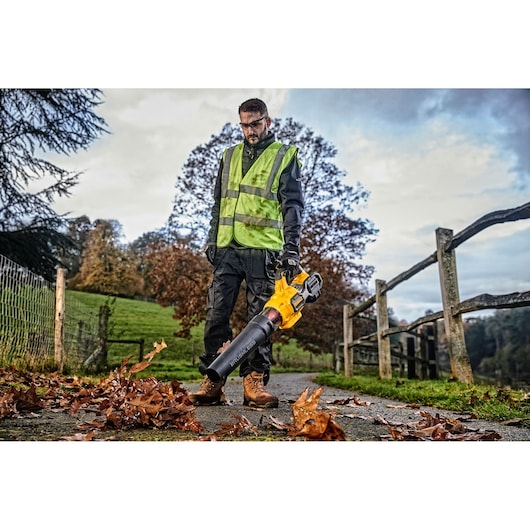 Person using a DEWALT leaf blower outdoors, wearing a safety vest and gloves, clearing leaves from a pathway.