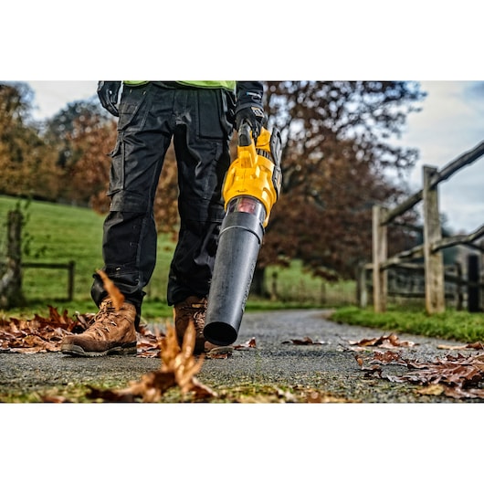 Person using a yellow DEWALT leaf blower on a paved path with scattered autumn leaves outdoors.