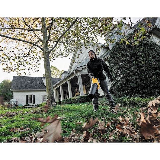 Person uses a DEWALT leaf blower to clear leaves in a yard outside a house.