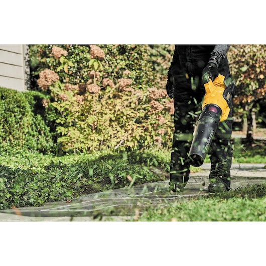 Person uses a yellow DEWALT leaf blower to clear grass clippings from a garden path outdoors.