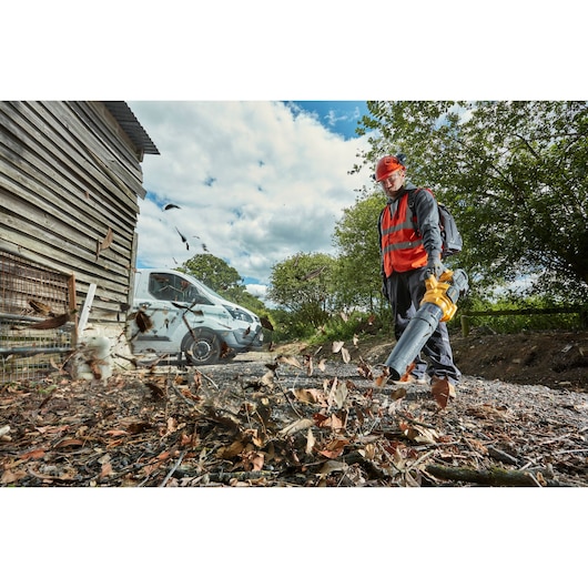 Person uses a DEWALT leaf blower outdoors, clearing leaves near a shed and van on a sunny day.